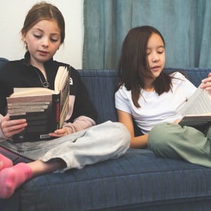 To jenter som sitter i en mørkeblå sofa og leser i hver sin bok 