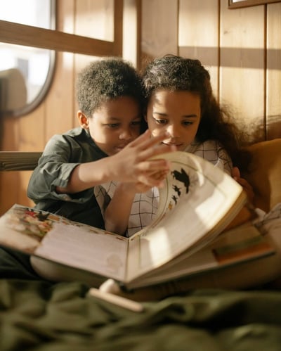 To barn som sitter under et pledd hjemme og blar i den samme boken. 